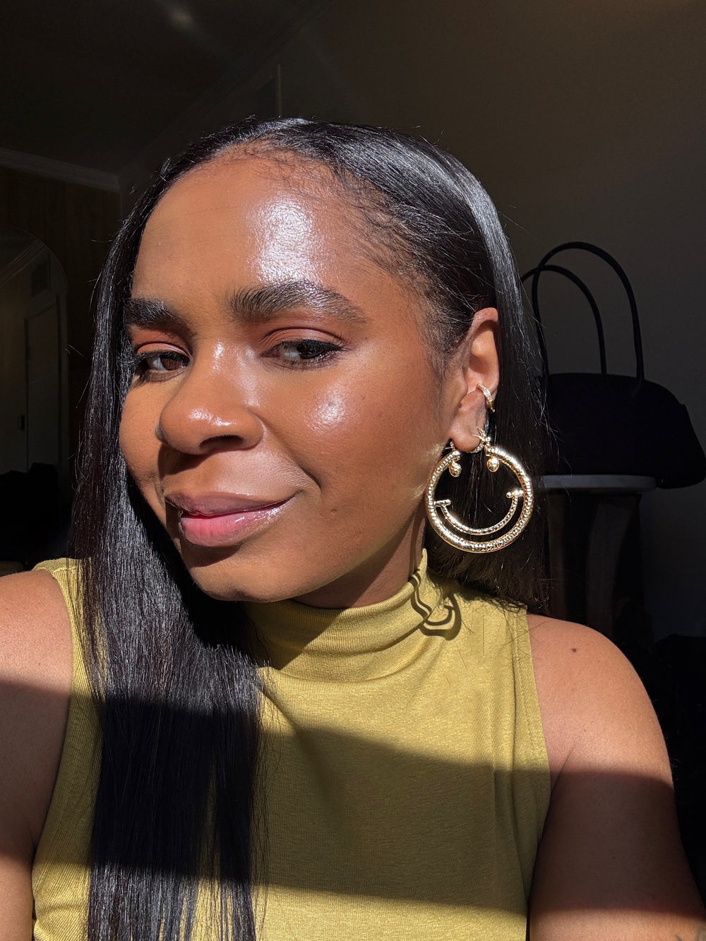 Woman with long straight hair wearing gold smiley face hoop earrings indoors.