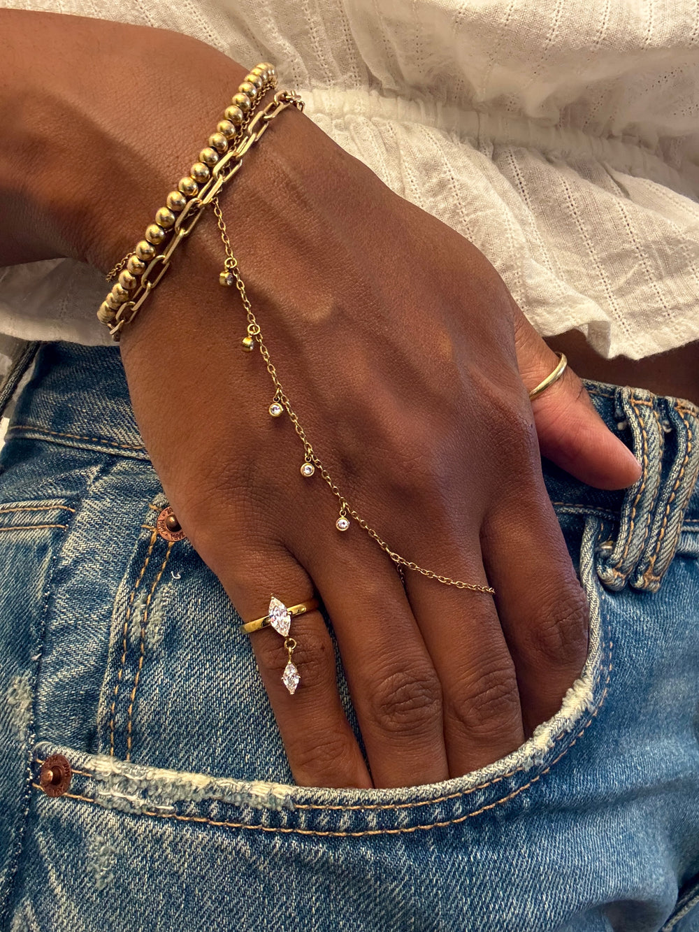 A close-up of a person's hand resting on blue denim jeans, showcasing a gold chain anklet with small clear stones draped over the wrist and hand, along with other gold jewelry.