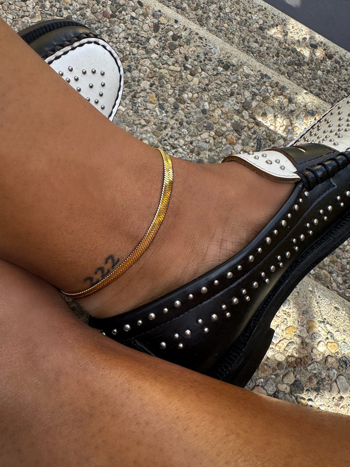 Black studded shoes worn on a foot with a gold anklet, on a textured pavement background.